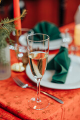 Close-up of a champagne flute on a red holiday table with green napkins, plates, and candlelight in the background. Elegant celebration vibe with copy space