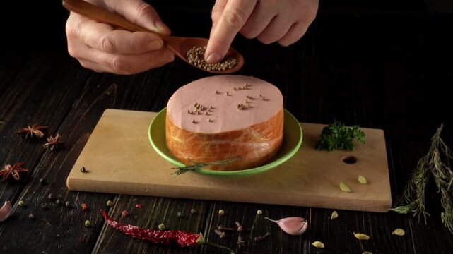 A person is preparing food by placing spices on a round piece of meat that sits on a wooden board. Herbs and various spices are scattered around the workspace.