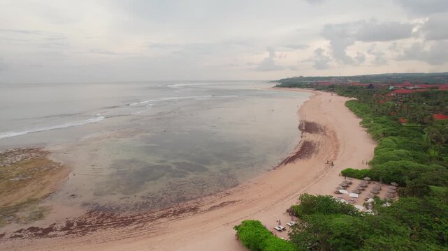 Aerial footage showcasing the sweeping curved shoreline of Geger Beach, Bali. Shallow waters glisten alongside lush greenery under soft, diffused light, creating a tranquil and scenic panorama.