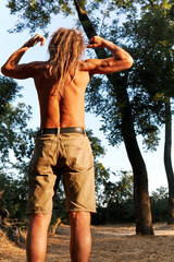 Shirtless man flexing muscles outdoors in warm sunset light surrounded by nature