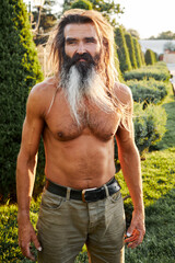 Shirtless mature man with long hair and beard standing outdoors in warm sunlight