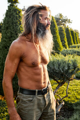 Shirtless mature man with long hair and beard standing outdoors in warm sunlight