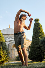 Shirtless bearded man stretching outdoors near historic building and evergreen trees