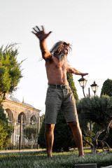 Shirtless bearded man stretching outdoors near historic building and evergreen trees