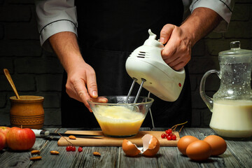 In a warmly lit kitchen, a person mixes batter using an electric mixer while surrounded by fresh ingredients like eggs, milk, and apples. The atmosphere feels homey and inviting