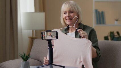 Old Caucasian woman recording video blog mobile phone on tripod showing pink jumper senior lady retired female influencer online trader show sweater clothes presenting review talking content creator