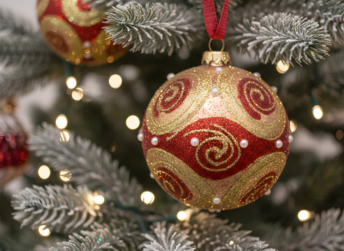 Close-up of a festive red and gold glitter Christmas ornament with pearls, hanging on a flocked fir branch with warm bokeh lights.