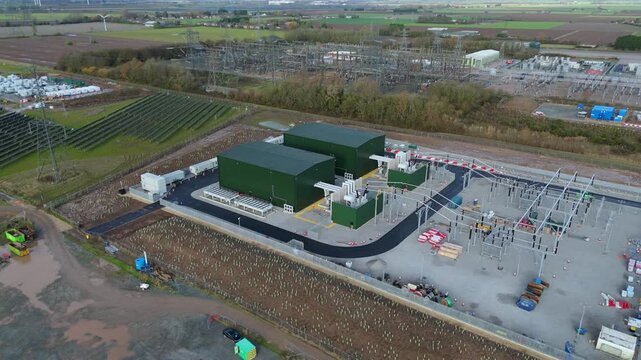 Aerial view of the power station with green buildings and high-voltage equipment surrounded by open fields and construction zones, Wisbech, England, United Kingdom.