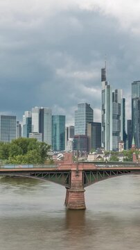 Aerial hyperlapse of Frankfurt skyline with Main River and Ignatz Bubis Bridge. Skyscrapers and church spires reflected in the water beneath dramatic clouds timelapse. Frankfurt, Germany