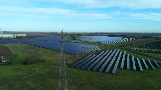 Aerial view of vast solar panel arrays contrasting with the green fields and a lattice transmission tower, creating a blend of modern and rural, Spalding, England, United Kingdom.