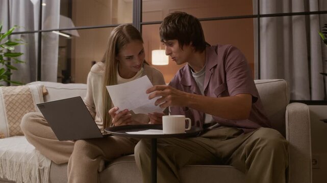 Medium shot of young Caucasian couple sitting on sofa in apartment, checking bills and managing finances on laptop, enjoying cozy and modern home environment