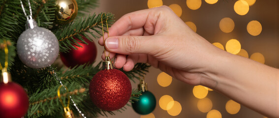 A close-up of a hand hanging a sparkling red ornament on a Christmas tree decorated with colorful baubles and warm bokeh lights, creating a cozy holiday atmosphere.