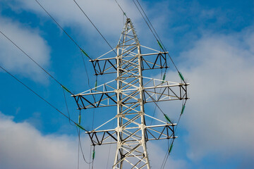 Striking power line pylon structure dominates the frame against a vibrant blue sky with scattered white clouds, showcasing industrial strength and overhead electricity transmission wires