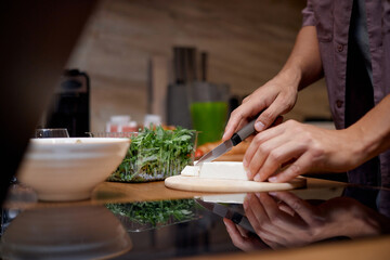 Obraz premium Caucasian young adult man slicing block of cheese on wooden cutting board in kitchen, hands visible holding knife, preparing food near fresh greens and salad ingredients
