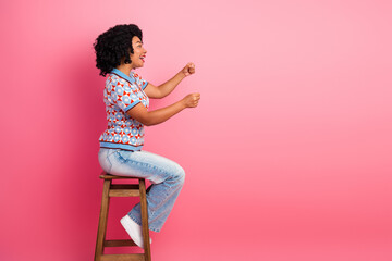 Happy young woman sits on a wooden stool in a pink studio wearing a stylish patterned shirt and jeans smiling with energy
