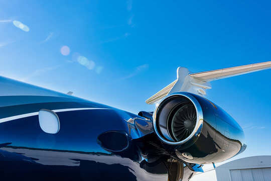 Close-up side view of a corporate jet showing the turbofan engine, fuselage, and, tail with visible lens flare and T-tail, taken in bright daylight under clear blue sky.