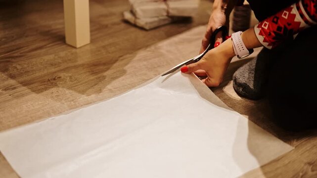 Closeup of hands cutting gift wrap on wooden floor during festive night indoors