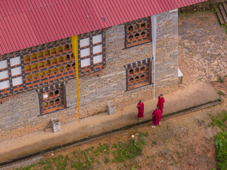 Aerial view of monks in vibrant red robes stand beside a stone building with intricate window details and a striking red roof, Paro, Bhutan.