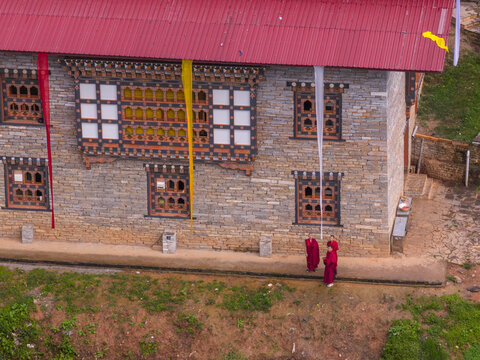 Aerial view of monks in vibrant red robes walking past a traditional Bhutanese building with intricate wooden window frames and colorful banners, Paro, Bhutan.
