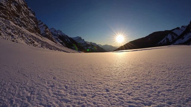 The sun rises slowly over a stunning, snow-covered landscape, casting a warm glow across the vast, white expanse. Majestic mountains tower in the distance, framing the tranquil sce