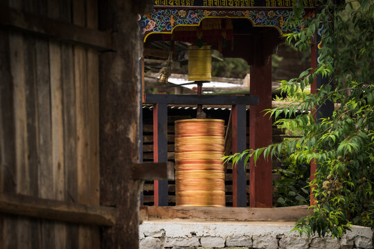 View of golden prayer wheels spinning gently in the dim light, framed by a weathered wooden door and lush greenery, Ogyencholing, Bumthang, Bhutan.