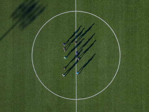 Aerial view of people standing on a bright green soccer field, their long shadows stretching dramatically within the circular boundary, Thimphu, Bhutan.