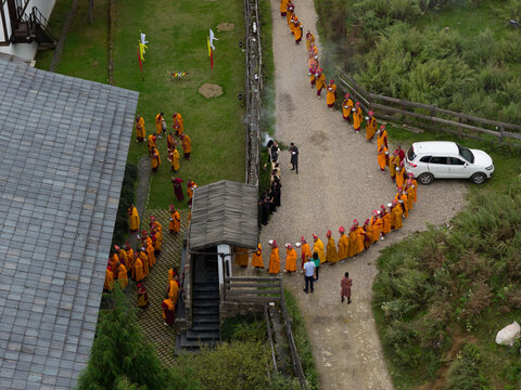 Gantey, Bhutan - 21 September 2025: Aerial view of monks in vibrant orange robes forming a procession along a path near the Gantey Monastery, contrasting with the green landscape and white vehicle.