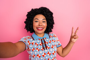 Smiling young woman in a geometric patterned shirt takes a pink studio selfie with a peace sign and cheerful vibe