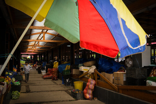 Trongsa, Bhutan - 20 September 2025: View of the market stalls under the vibrant umbrellas, casting colorful shadows across the stone floor, as a lone figure walks by stacks of produce.