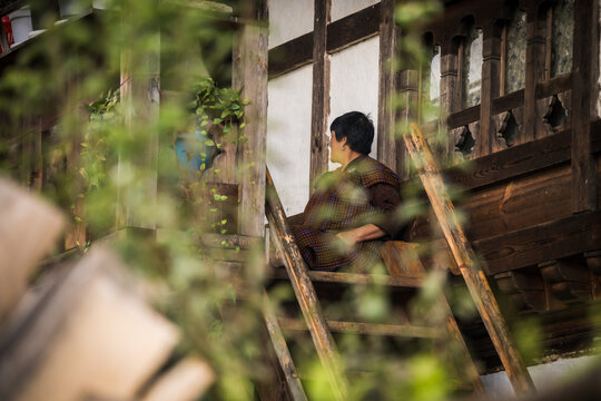 Ogyencholing, Bhutan - 23 September 2025: View of a person seated on the wooden stairs of a traditional building, framed by vibrant green foliage, evoking a sense of serene contemplation.