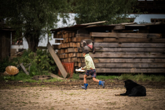 Ogyencholing, Bhutan - 23 September 2025: View of a boy running across the dusty ground with a tray, as a black dog watches amidst stacked wood and sparse greenery.