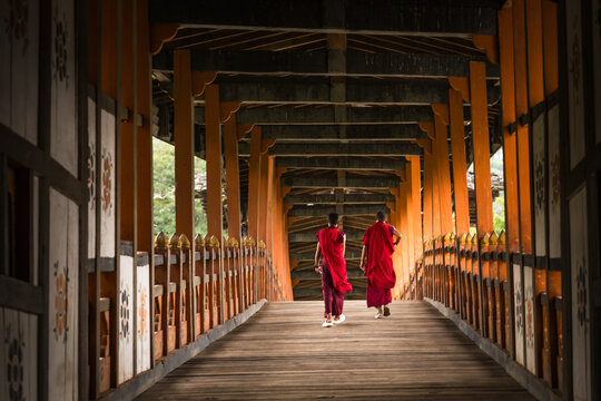 Punakha, Bhutan - 19 September 2025: View of two monks in vibrant red robes walking across a wooden bridge, its orange beams casting shadows, a serene journey captured in time.