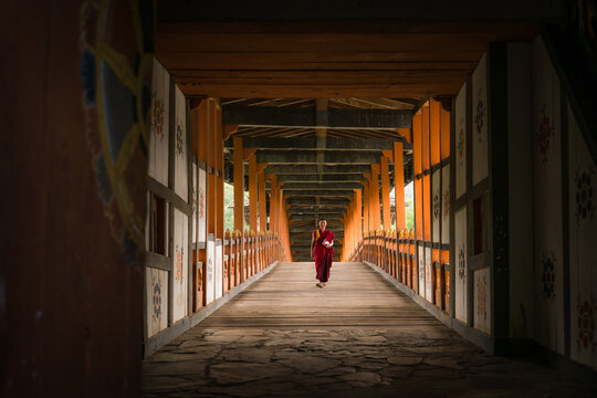 Punakha, Bhutan - 19 September 2025: View of a lone monk in vibrant red robes walking across a wooden bridge, the light filtering through the structure's intricate frame.
