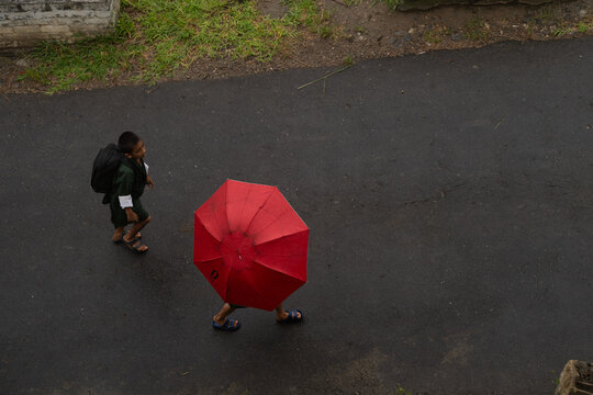 Punakha, Bhutan - 19 September 2025: View of a lone boy in school uniform and backpack, shadowed by a vibrant red umbrella against the dark, glistening asphalt, strides through the quiet city street.