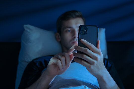 Sleepless young man browsing on his phone, doom scrolling in bed late at night lit by glowing blue screen light. Insomnia, procrastination or social media addiction. Selective focus on smartphone