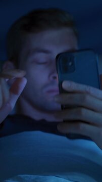 Late at night, tired man lying awake in bed, stares at his glowing mobile phone, doom scrolling social media out of habit before reacting with visible frustration from screen addiction and insomnia. 