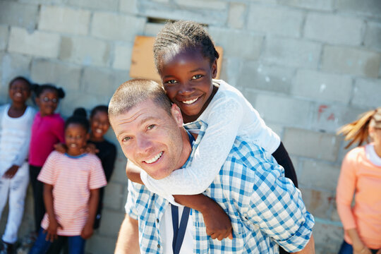 Portrait, piggyback and man with girl, community outreach and children with support. Adoption, people and volunteer with orphan kids, smile and happiness with charity event, playing and kindness