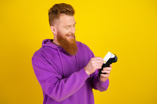 Young bearded man in purple hoodie uses a card reader on a bright yellow background for shopping and advertising