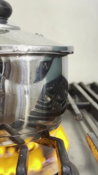 Vertical video social media - Closeup of a lid being removed from an overfilled stainless steel pan of boiling water on a gas cooker hob, which is spilling out over the sides into an orange flame.