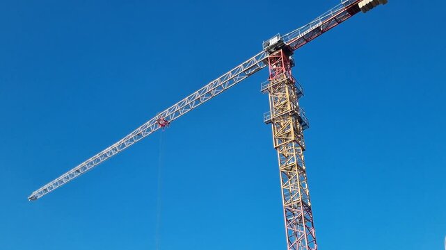Tall construction tower crane rotating its jib arm against a clear blue sky. Heavy lifting machinery and equipment at a building site