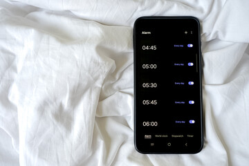 Overhead view of a smartphone displaying multiple alarms on a rumpled white sheet or blanket.