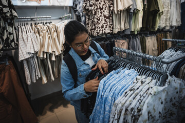 Fototapeta premium Indian woman browsing clothes inside a modern fashion retail store, selecting garments from a clothing rack.