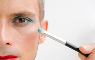 portrait of a feminine gay man with makeup and cosmetics on face. Transgender male with red lipstick applied eye shadow sparkle on his eyes with a brush on a white background