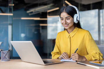 Smiling young Indian woman wearing headphones sitting at her desk, looking at her laptop screen and...