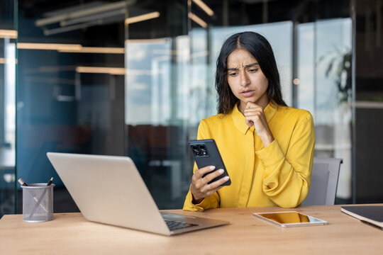 Worried young Muslim businesswoman sitting at a desk in the office and looking at her mobile phone screen in frustration