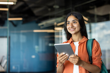 Close-up photo of a young Indian female student standing indoors with a backpack, holding a tablet in her hand and looking ahead