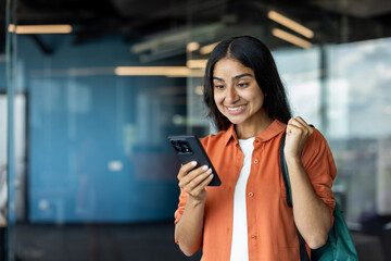 A young Jewish female student is standing in a room, looking at the screen of her mobile phone and being happy