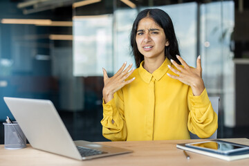 Worried young Indian woman sitting in office feeling very hot, fanning herself with her hands