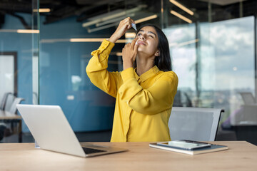Tired young Muslim woman working in the office, sitting at her desk and putting eye drops in her eyes