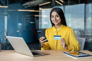 Portrait of a smiling young Muslim woman sitting at a desk in the office, using a phone and credit card, looking at the camera